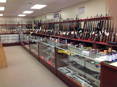 A gun store interior with rifles displayed on wall racks and various ammunition and accessories in glass display cases below.